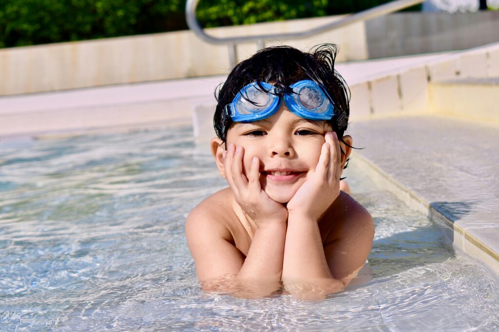 A young child with goggles smiling while playing in an outdoor swimming pool.